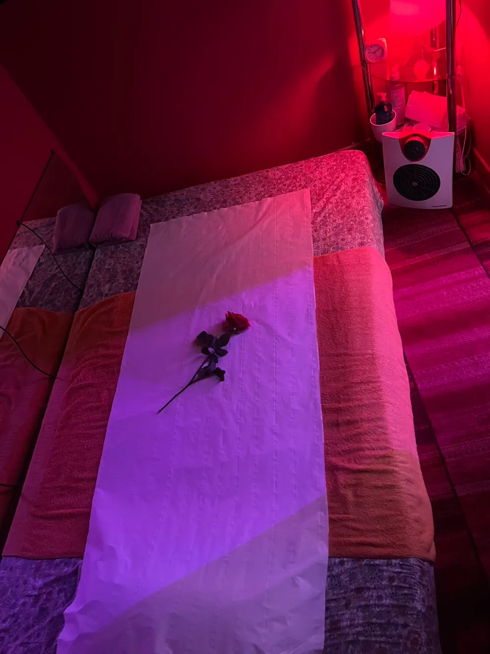 Vue d’en haut de la table de massage : un drap blanc et une rose rouge, sous la lampe rouge de la cabine